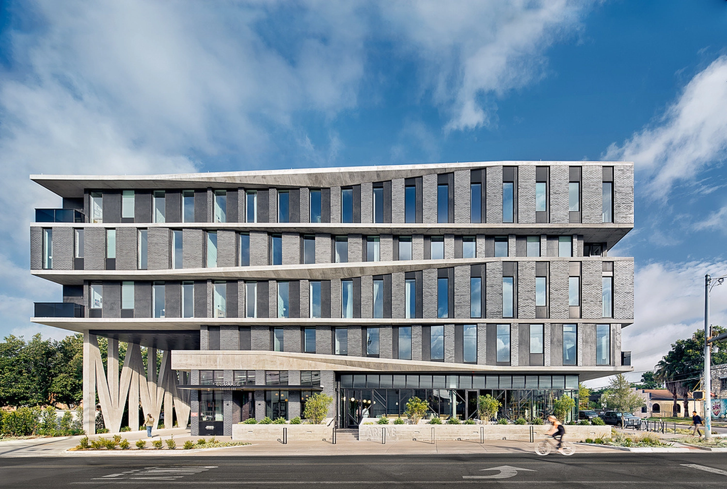 Izudin Jasarevic – Pflege mit Menschlichkeit – Eine Ansicht des ARRIVE Austin Hotels aus der Froschperspektive. Die moderne Fassade besteht aus grauen Ziegeln und markanten, kantigen Betonbändern oder -vordächern. Das Gebäude verfügt über mehrere Reihen vertikaler Fenster, ein Erdgeschoss mit verglasten Schaufenstern und einen klaren blauen Himmel mit leichten Wolken im Hintergrund. Im unteren rechten Bildbereich ist eine Person auf einem Fahrrad teilweise zu erkennen.