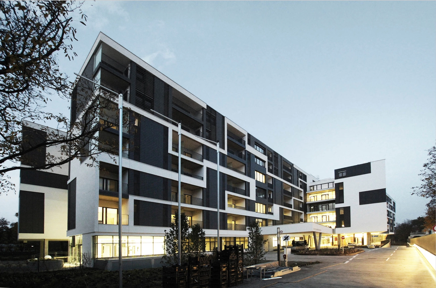 Izudin Jasarevic – Pflege mit Menschlichkeit – Ein modernes, mehrgeschossiges Wohngebäude in der Abenddämmerung mit einer markanten geometrischen Fassade aus dunkelgrauen und weißen rechteckigen Paneelen und Balkonen. Große Glasfenster im Erdgeschoss werden von innen hell erleuchtet, während sich ein auskragendes, weißes Eingangsvordach über die Zufahrt erstreckt. Schmale, hohe Lichtmasten ragen im Vordergrund vor dem klaren Abendhimmel empor.