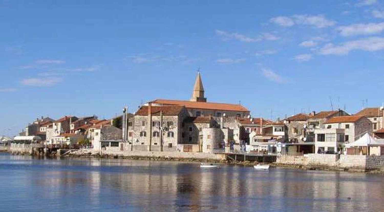 Izudin Jasarevic - Pflege mit Menschlichkeit - Panoramablick auf die historische Altstadt von Umag, Kroatien, mit Steingebäuden mit Terrakotta-Dächern und einem markanten Kirchturm entlang des blauen Wassers der Adriaküste unter klarem Himmel.