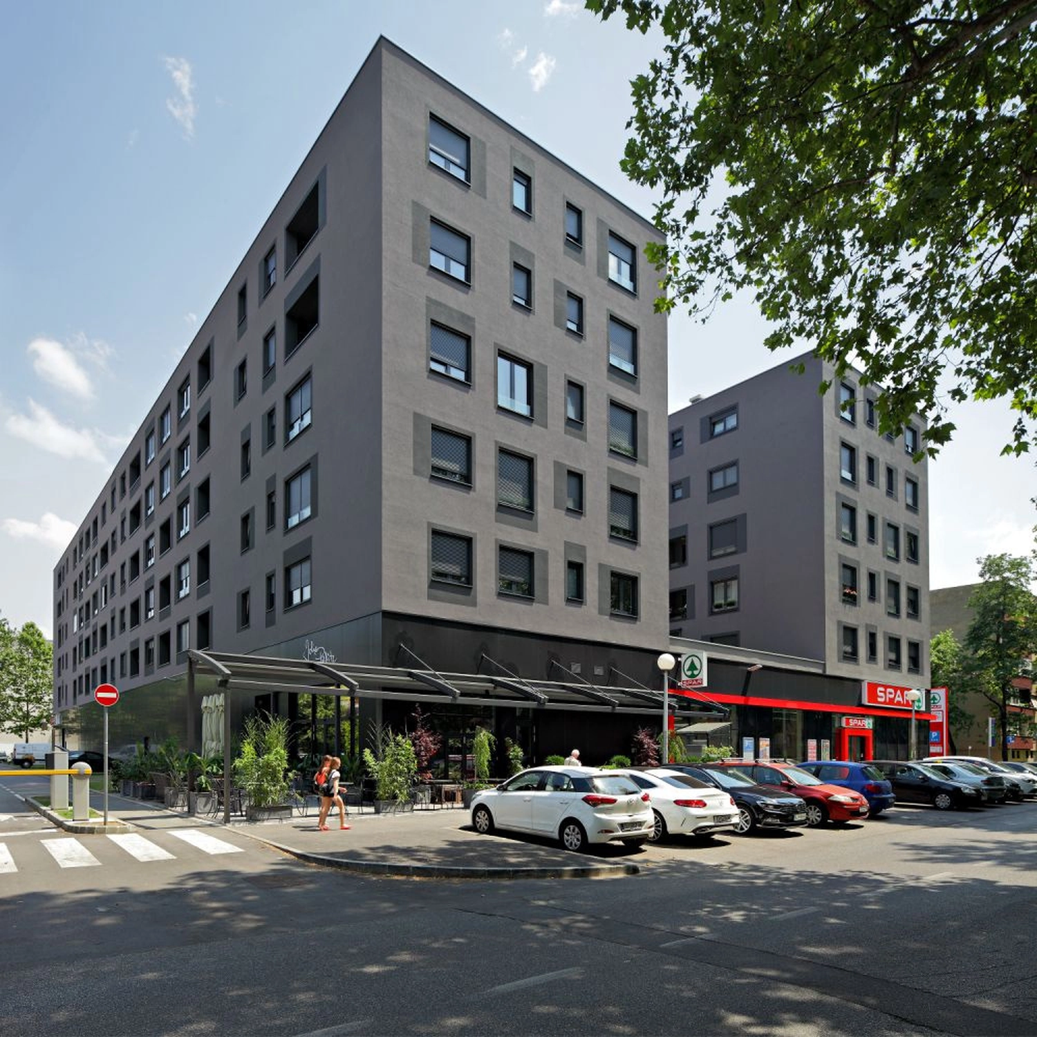 Izudin Jasarevic – Pflege mit Menschlichkeit – Das Bild zeigt einen großen, modernen, grauen Apartmentkomplex mit mehreren Stockwerken und Flachdach. Im Erdgeschoss befindet sich ein SPAR-Supermarkt, erkennbar an seinem roten Branding und Logo. Vor dem Gebäude liegt ein Parkplatz mit mehreren Autos (weiß, rot und schwarz) und ein Gehweg, auf dem Fußgänger unterwegs sind. Ein Baum mit grünem Laub rahmt die obere rechte Ecke des Bildes ein. Die Architektur zeichnet sich durch klare Linien, rechteckige Fenster mit dunklen Rahmen und einen dunklen Sockel aus.