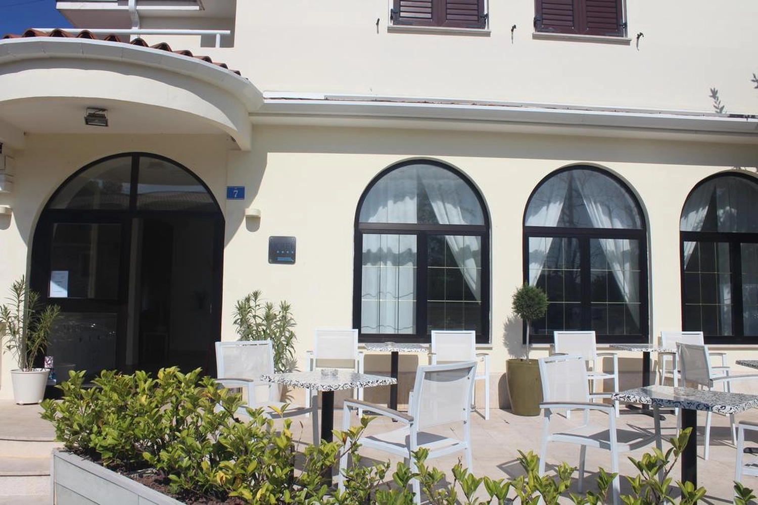 Izudin Jasarevic – Pflege mit Menschlichkeit – Außenansicht der weißen Fassade des Hotels La Settima Luna mit einer Außenterrasse, die mit modernen weißen Stühlen und Marmortischen ausgestattet ist. Das Gebäude verfügt über mehrere große, schwarz gerahmte Bogenfenster mit weißen Vorhängen und einen Haupteingang mit einer ähnlichen Bogentür aus Glas. Kleine grüne Topfpflanzen stehen im hellen Tageslicht in der Nähe des Eingangs und auf der Terrasse.