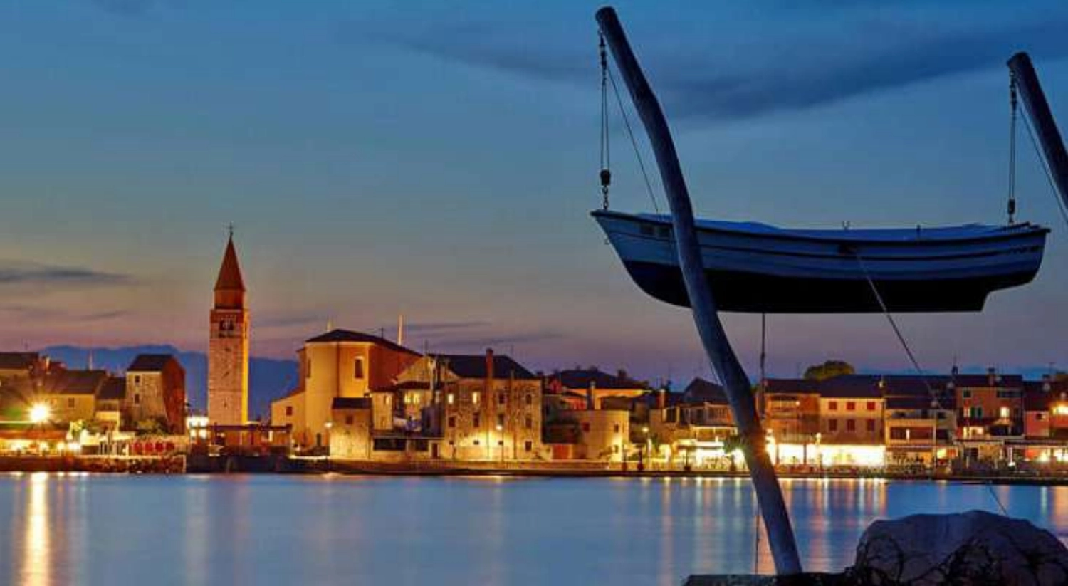 Izudin Jasarevic – Pflege mit Menschlichkeit – Ein malerischer Blick in die Abenddämmerung auf die historische Stadt Umag in Kroatien. Im Vordergrund sind der markante Kirchturm und die alten, beleuchteten Steingebäude entlang der Uferpromenade zu sehen. Ein traditionelles Holzboot hängt an einem Kran oder Hebezeug und zeichnet sich vor dem tiefer werdenden blauen Himmel und den Lichtreflexen im ruhigen Hafenwasser ab.